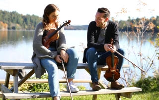 Jane Cory and Kyle Burghout with fiddles sitting by a lake
