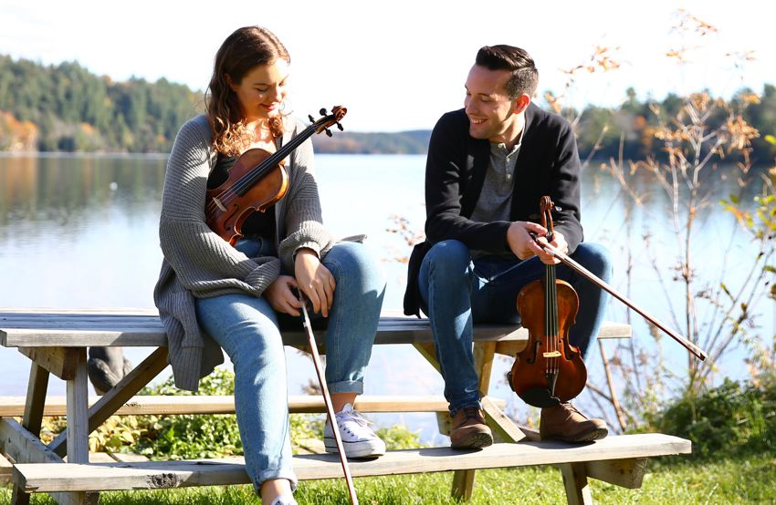 Jane Cory and Kyle Burghout with fiddles sitting by a lake
